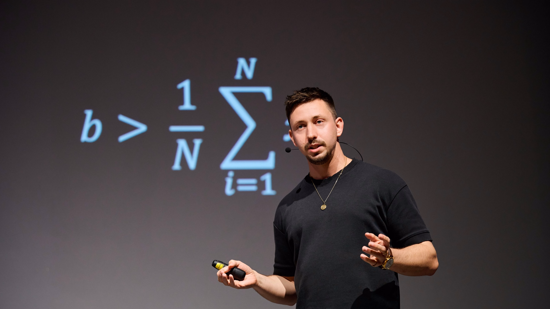 Max Fröhlich spricht mit Headset-Mikrofon und Presenter in der Hand auf einer Bühne, hinter ihm eine projizierte Formel mit Summenzeichen.