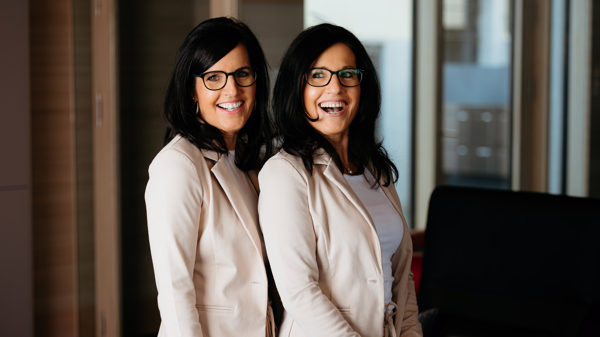 Regina Diez und Claudia Thum stehen in beigen Blazern nebeneinander vor einer Fensterfront und lachen.