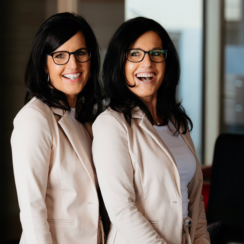 Regina Diez und Claudia Thum stehen in beigen Blazern nebeneinander vor einer Fensterfront und lachen.