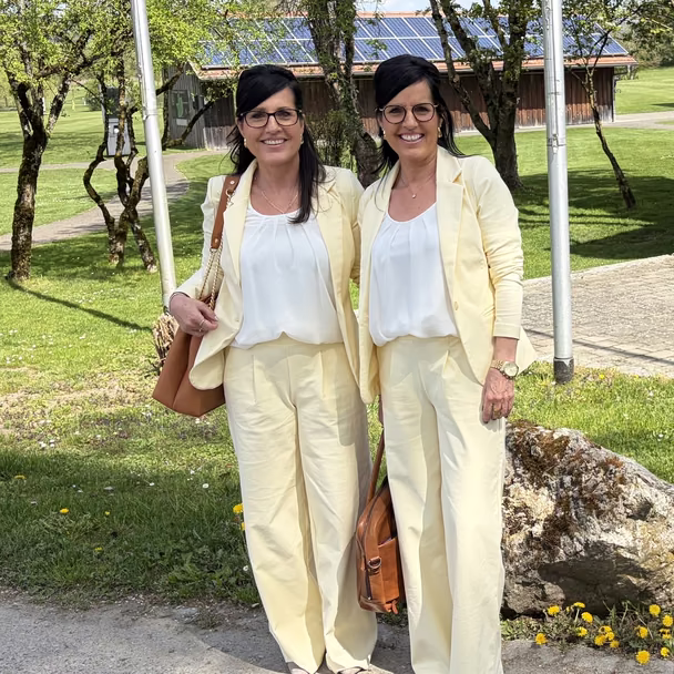 Regina Diez und Claudia Thum in hellgelben Anzügen auf einer Wiese vor Bäumen und einer Scheune mit Solardach.