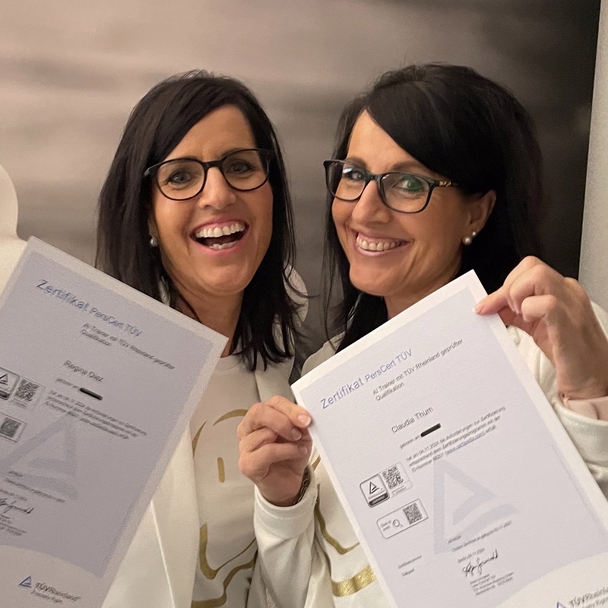 Regina Diez und Claudia Thum halten lachend ihre TÜV-Zertifikate als KI-Trainerinnen in die Kamera.