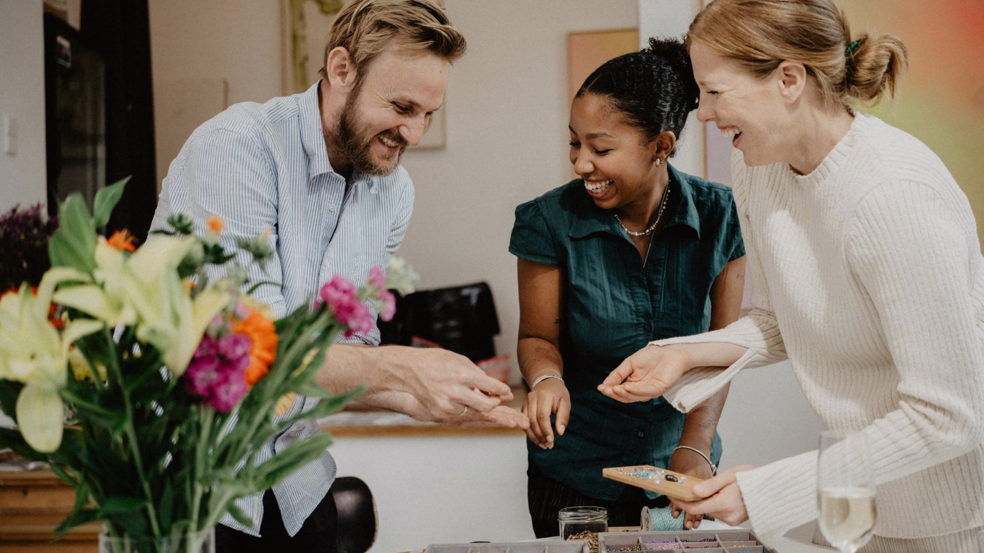 Jasper Kolb und zwei Frauen lachen an einem Tisch mit Perlen und Garnrollen, im Vordergrund ein bunter Blumenstrauß.