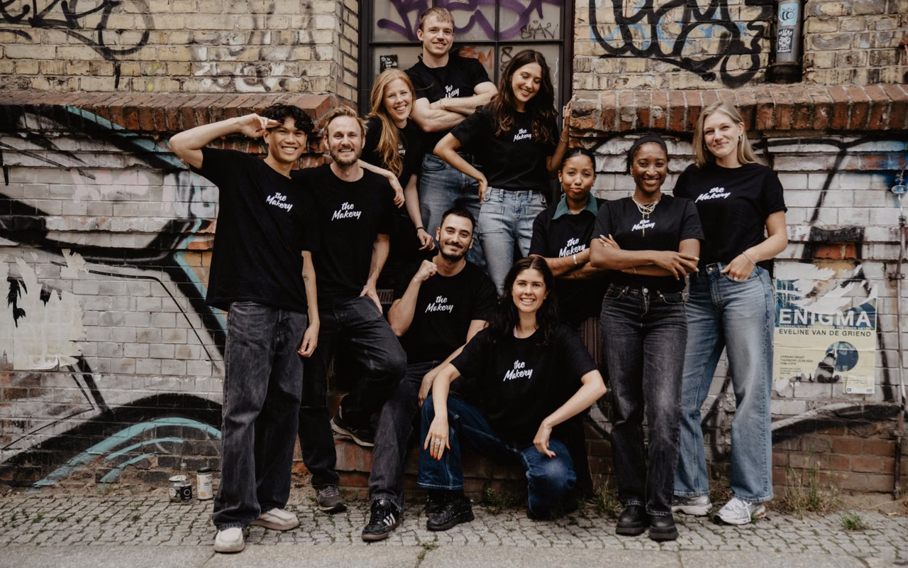 Das Team von The Makery in schwarzen T-Shirts mit Logo posiert vor einer Backsteinwand mit Graffiti.