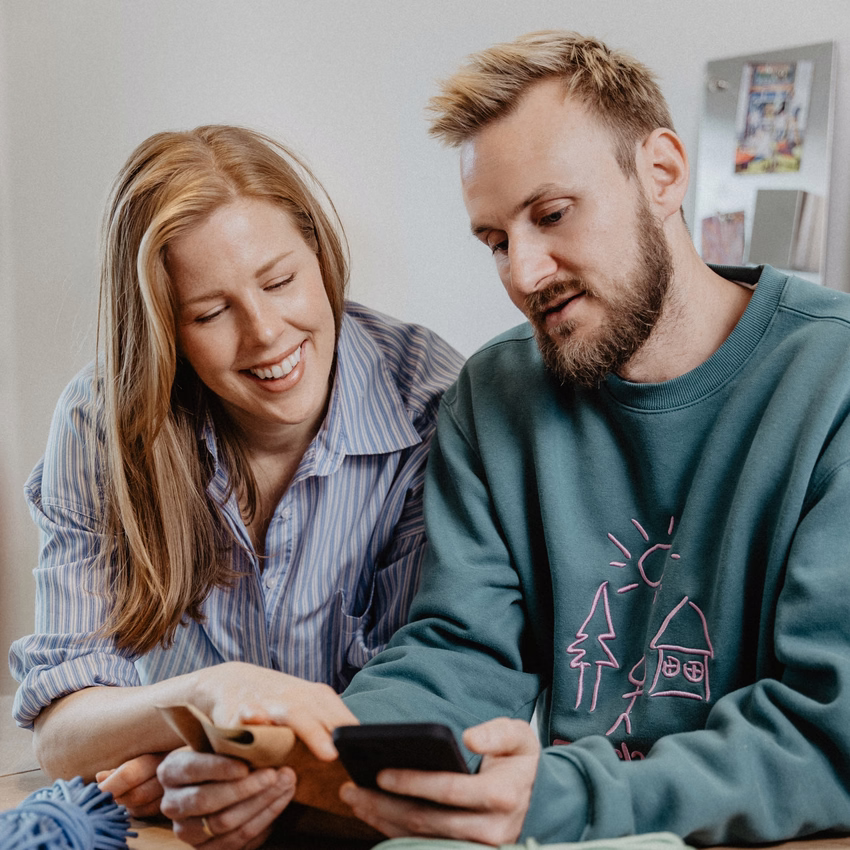 Jasper Kolb und eine Frau schauen lächelnd auf ein Smartphone, vor ihnen auf dem Tisch liegen Kordeln und ein Maßband.