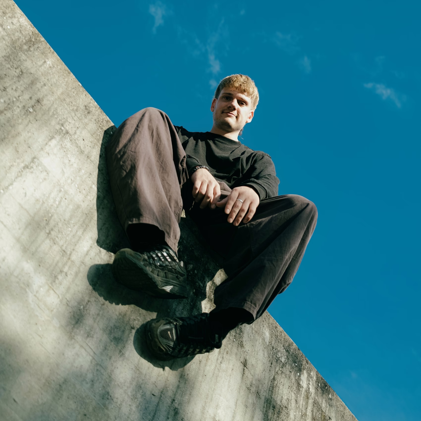 Martin Meyer sitzt lässig auf einer schrägen Betonmauer vor blauem Himmel, von unten fotografiert.