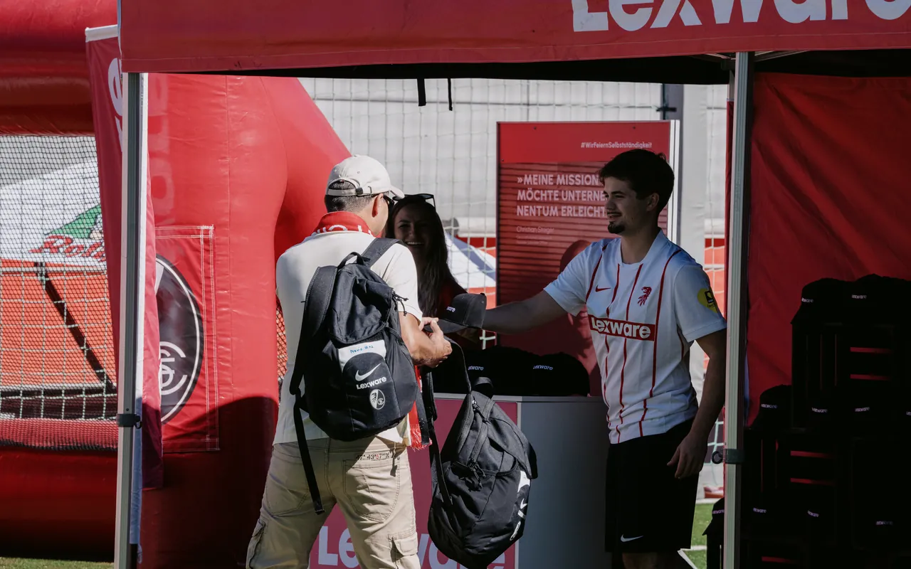 Mitarbeiter im Lexware-Trikot reicht einem Besucher am roten Lexware-Stand eine Cap über den Tresen.