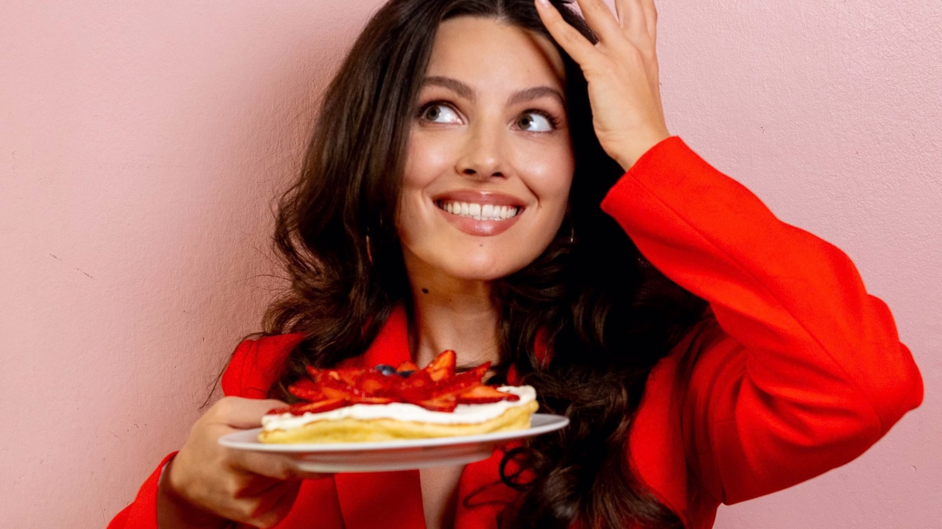 Leoni Lencinas im roten Blazer hält lachend einen Teller mit Erdbeer-Dessert vor einer rosa Wand, die andere Hand im Haar.