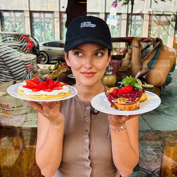 Eine Frau mit schwarzer Kappe hält zwei Teller mit Frühstücksdesserts in die Kamera, hinter ihr die Straße.