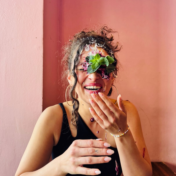 Eine lachende Frau, das Gesicht teils mit Blüten und Kräutern bedeckt, hält sich die Hand vor den Mund.