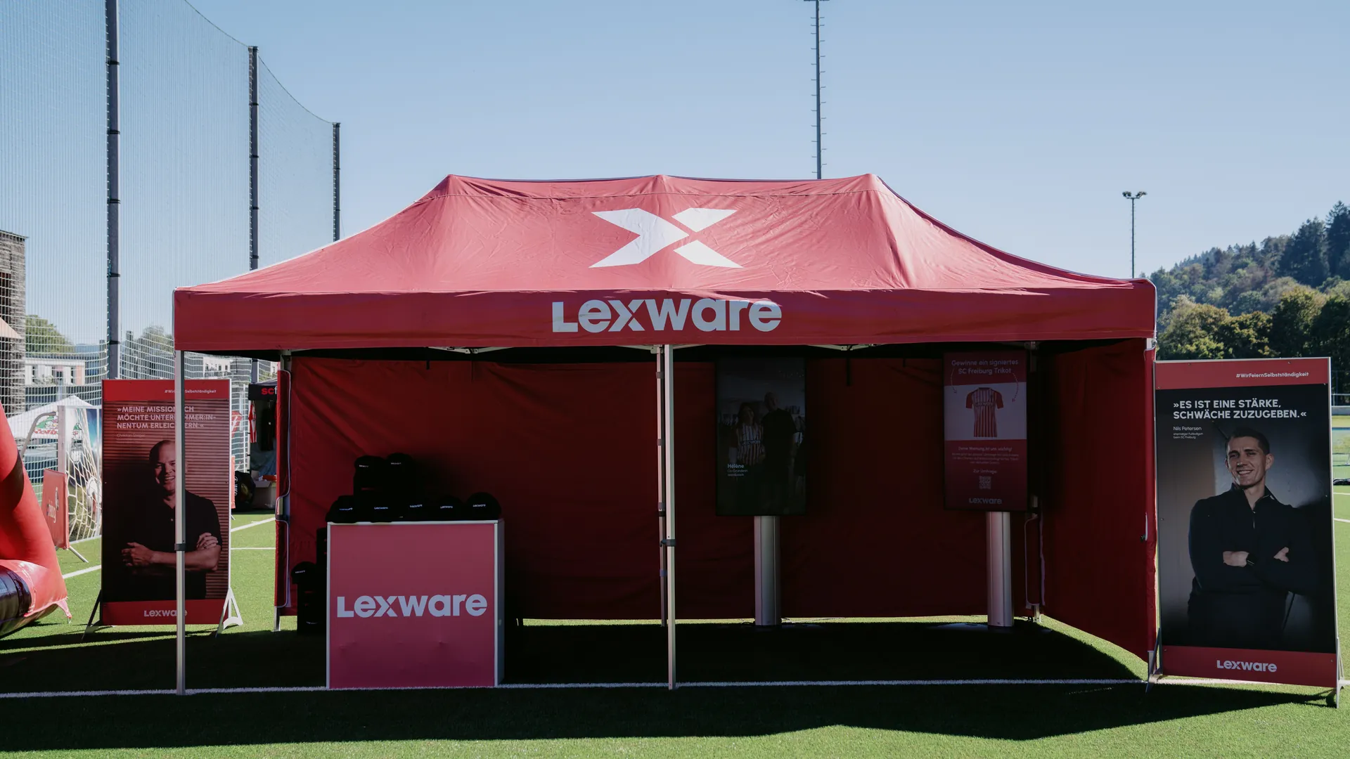 Roter Lexware-Pavillon mit Logo auf dem Dach, davor Aufsteller und ein Tresen, dahinter bewaldete Hügel unter blauem Himmel.