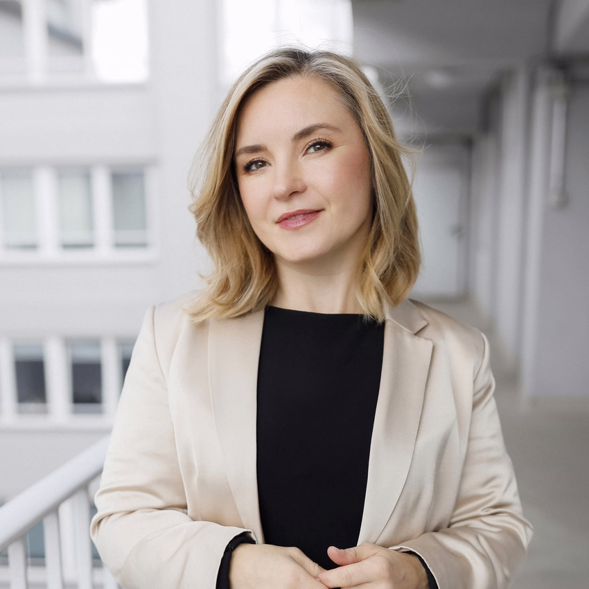 Elena Bärweg in hellem Blazer steht auf einem Laubengang vor einer weißen Hausfassade.