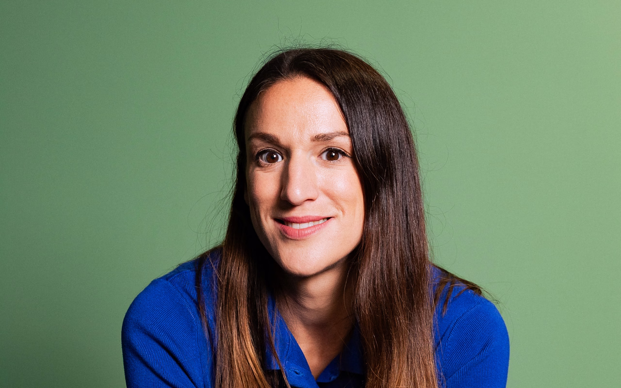 Caroline Nichols im blauen Poloshirt lächelt vor einer grünen Wand in die Kamera.