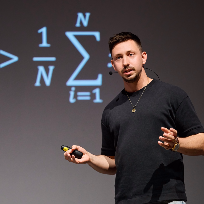 Max Fröhlich spricht mit Headset-Mikrofon und Presenter in der Hand auf einer Bühne, hinter ihm eine projizierte Formel mit Summenzeichen.