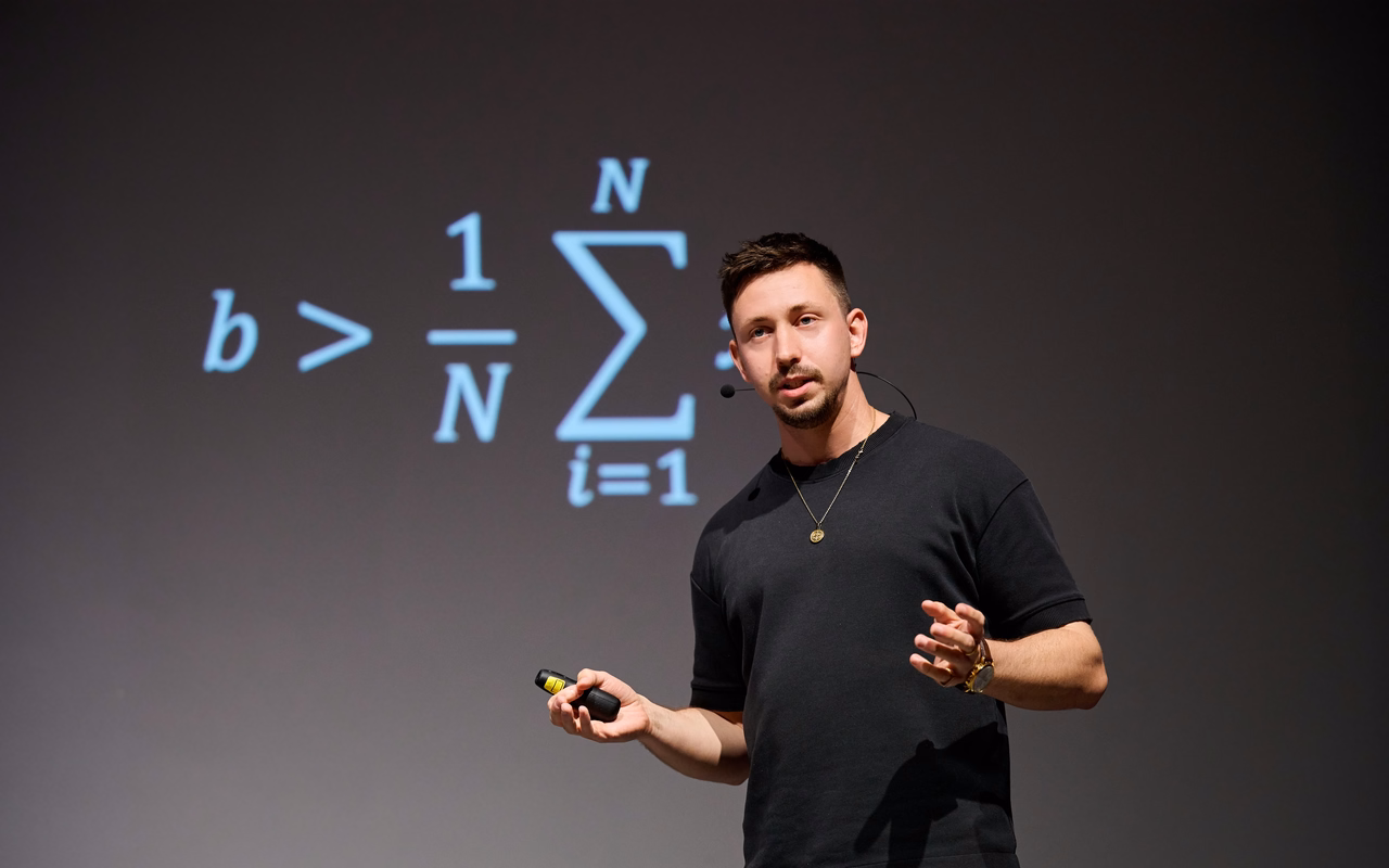 Max Fröhlich spricht mit Headset-Mikrofon und Presenter in der Hand auf einer Bühne, hinter ihm eine projizierte Formel mit Summenzeichen.