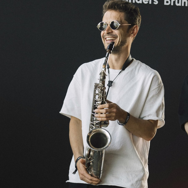 Kamil Khusyainov steht lachend mit Saxophon und Sonnenbrille vor einer dunklen Wand.