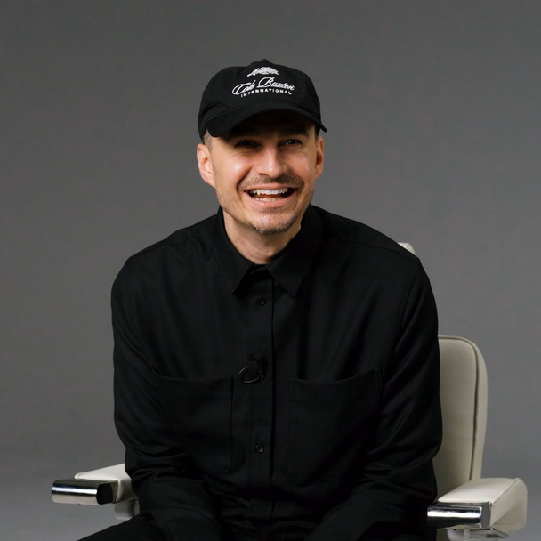 Christoph Filser sitzt lachend in schwarzer Kleidung und Basecap auf einem weißen Friseurstuhl vor grauem Studiohintergrund.