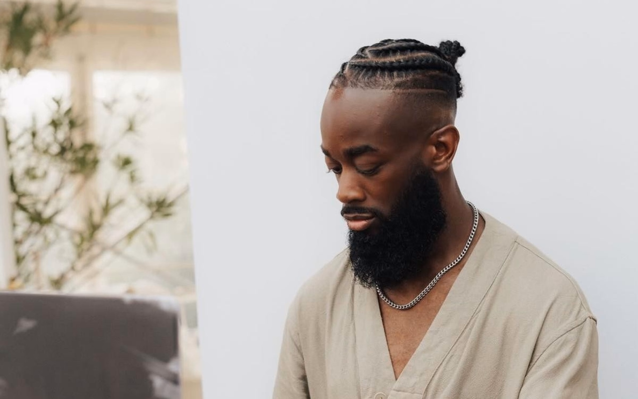 Noel Tagoe in hellem Hemd mit Silberkette steht am DJ-Pult und schaut nach unten, links unscharf Pflanzen vor einem Fenster.
