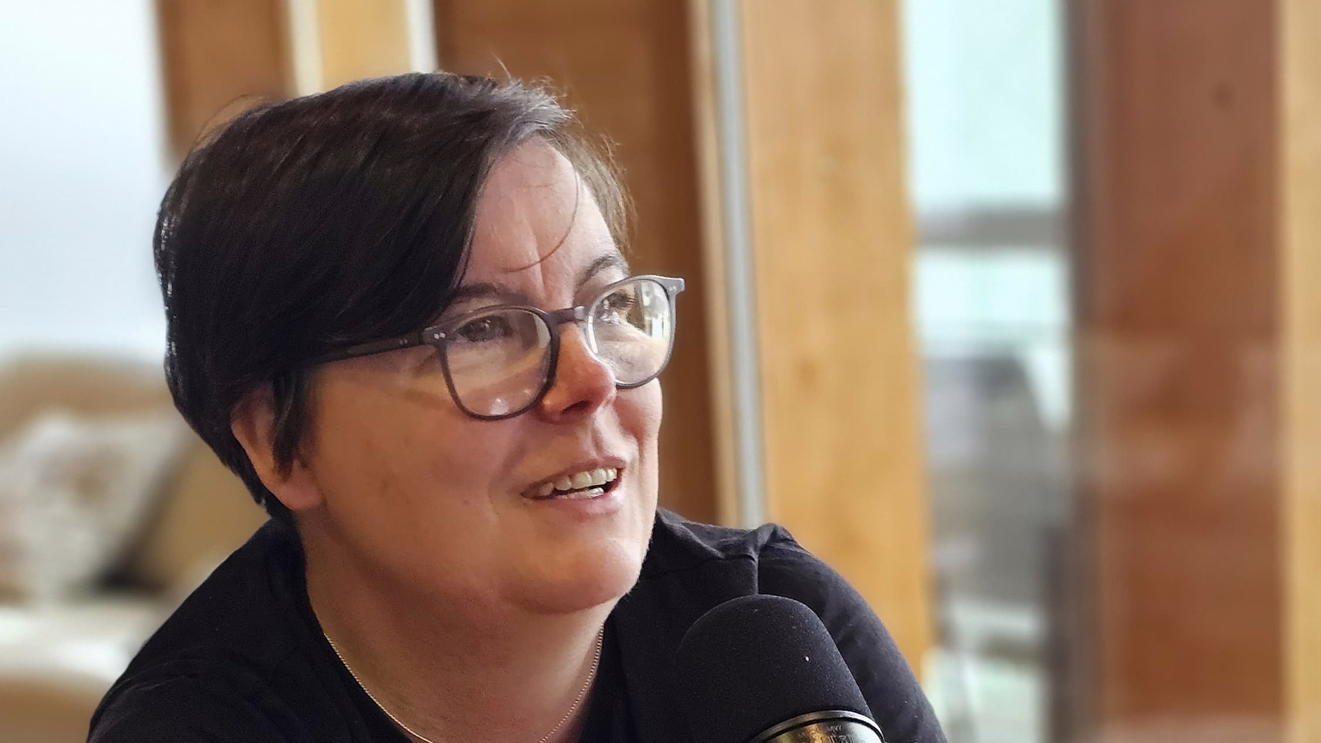 Britta Behrens mit Brille spricht lächelnd in ein Mikrofon, im Hintergrund Holz und Fenster.