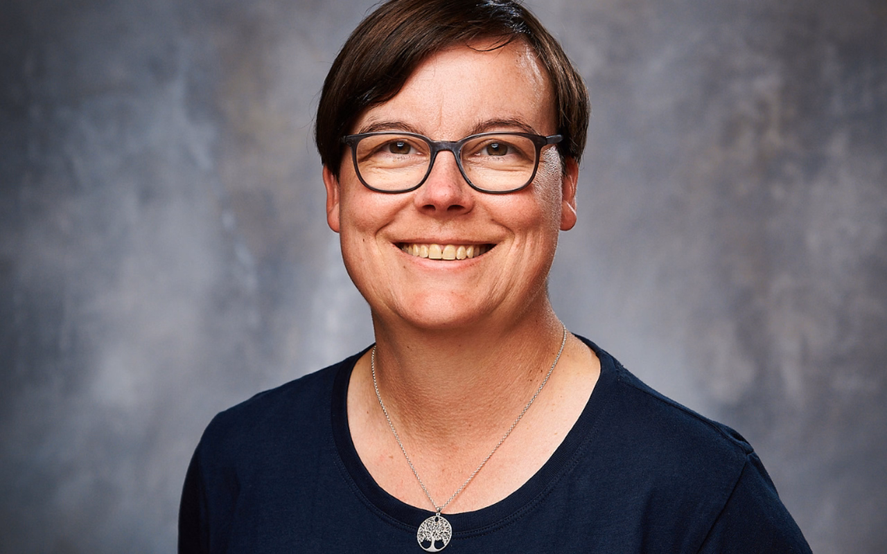 Porträt von Britta Behrens mit Brille und dunklem T-Shirt vor grauer Studiowand.