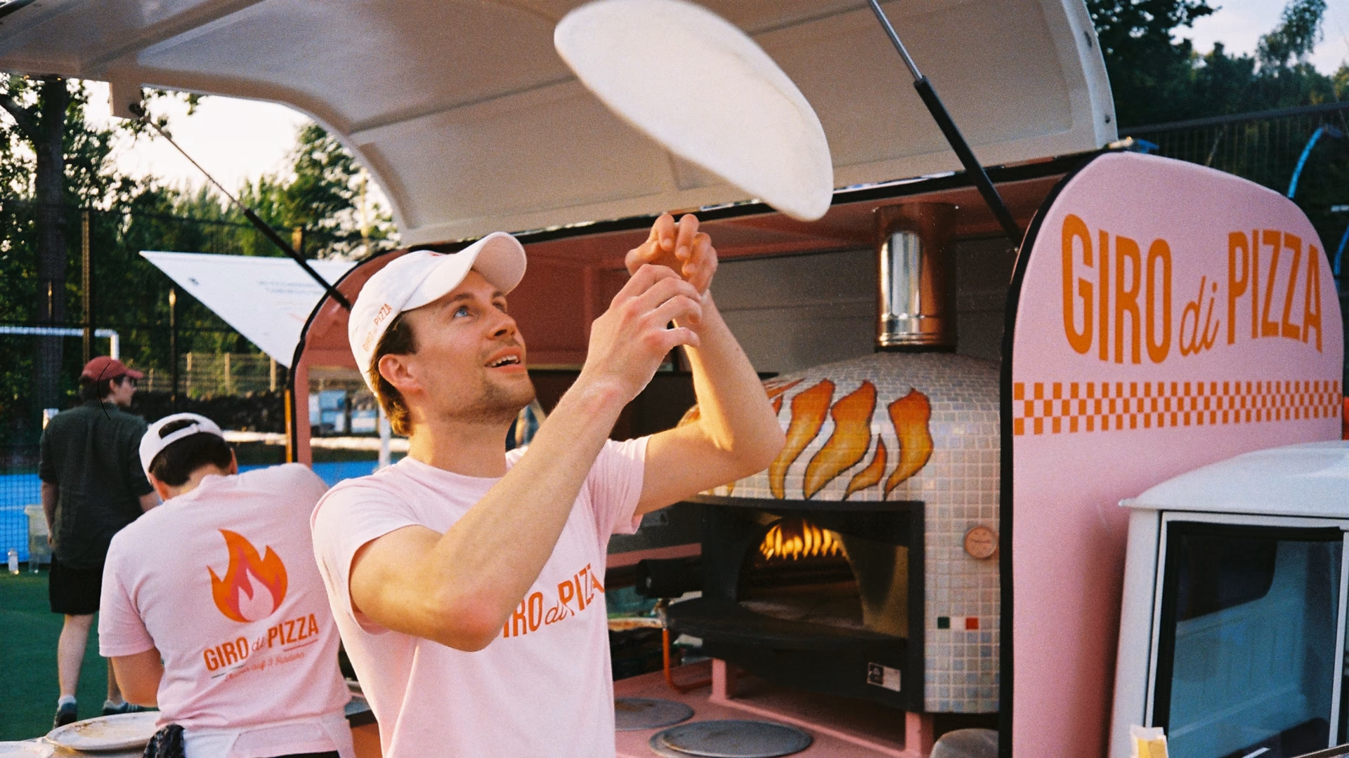 Anton Calamita wirft vor seiner rosa Pizza-Ape einen Pizzateig in die Luft, dahinter der Ofen mit Flammenmosaik und der Schriftzug Giro di Pizza.