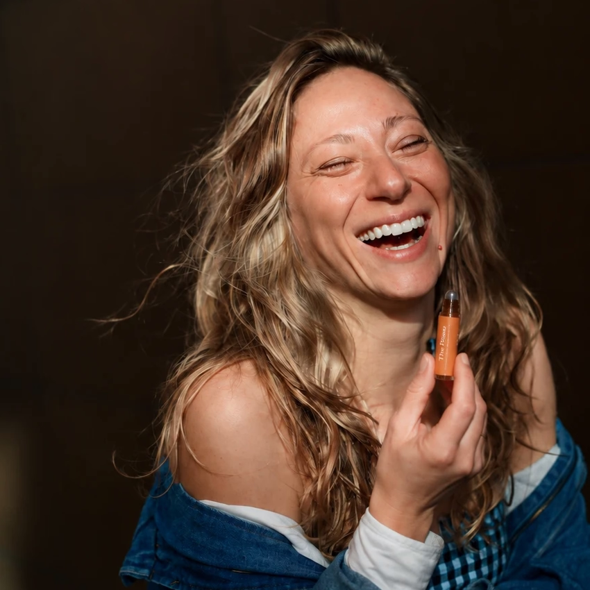 Elena Margulis lacht und hält einen orangefarbenen Duft-Roll-on neben ihr Gesicht.
