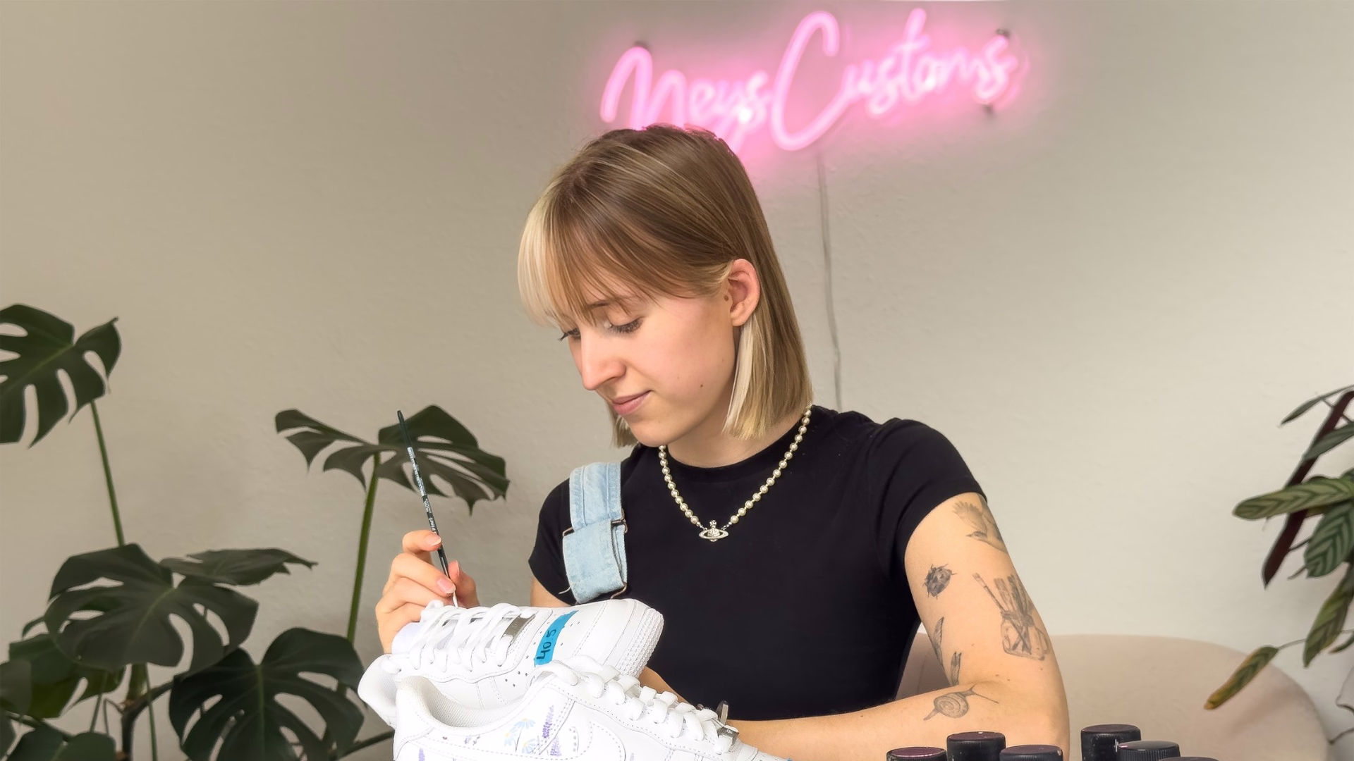 Nele Burgstaler bemalt mit einem feinen Pinsel einen weißen Sneaker, hinter ihr leuchtet der pinke Schriftzug NeysCustoms an der Wand.