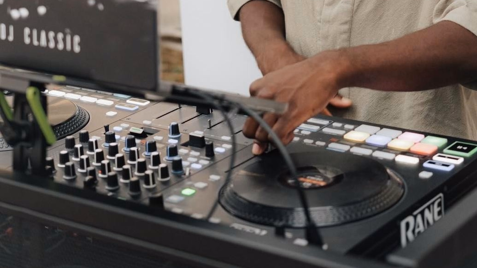 Noel Tagoes Hand mischt am DJ-Pult mit leuchtenden Pads, links ein Laptop mit der Aufschrift „DJ Classic“.