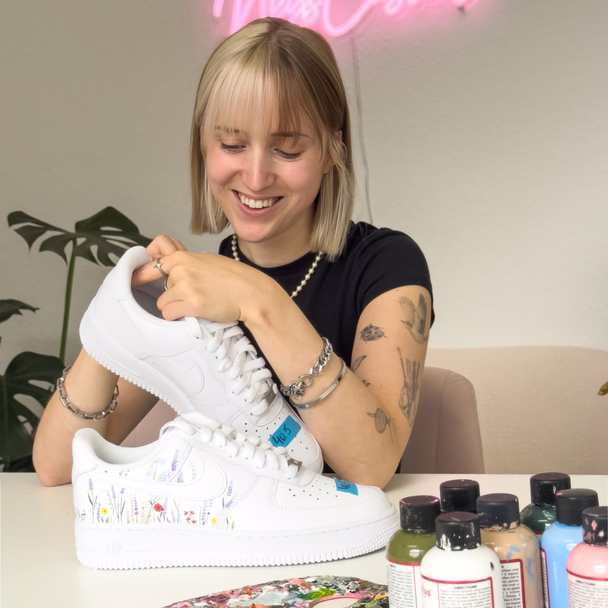 Nele Burgstaler hält einen weißen Sneaker mit gemalten Wildblumen in die Kamera, auf dem Tisch stehen Farbflaschen.