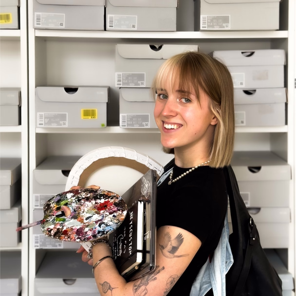 Nele Burgstaler steht mit Farbpalette und Notizbuch vor einem Regal voller Schuhkartons.