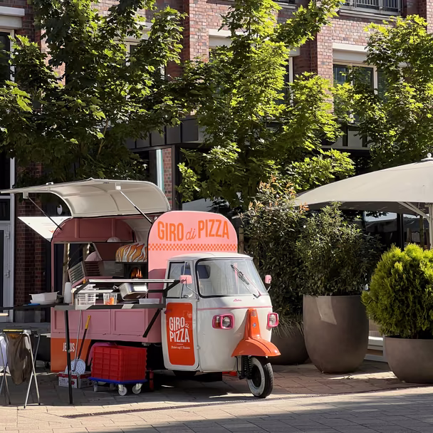 Die rosa-weiße Piaggio Ape von Giro di Pizza mit aufgeklapptem Verkaufsdach vor einer Backsteinfassade.