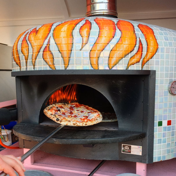 Eine Pizza auf der Schaufel vor dem Pizzaofen mit Flammenmosaik in der Ape.