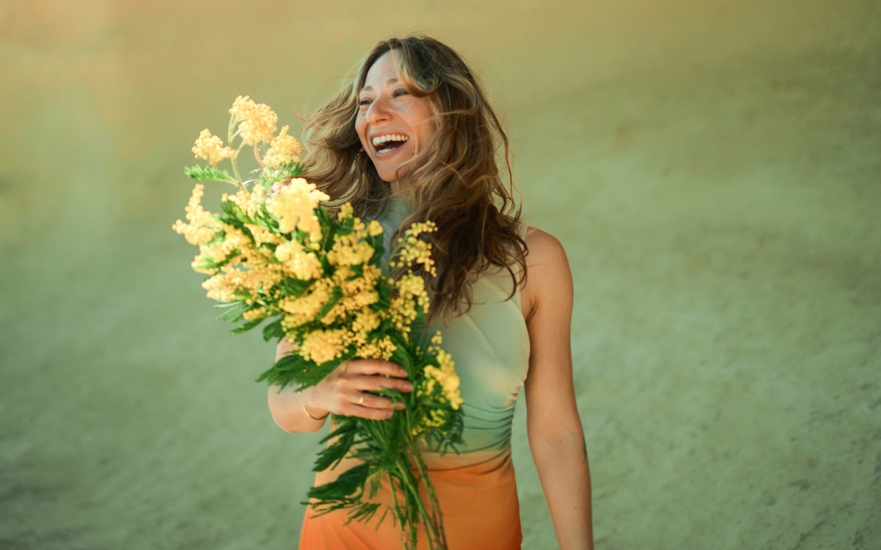 Elena Margulis lacht mit einem großen Strauß gelber Mimosen im Arm, hinter ihr eine sandfarbene Wand.