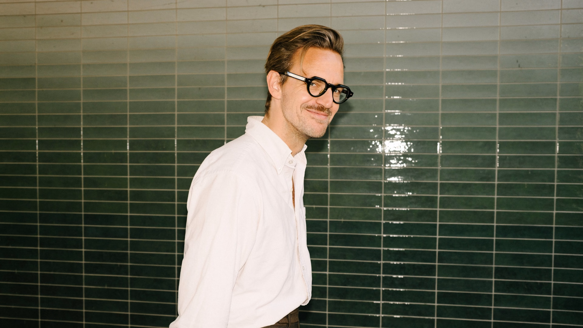 Sebastian Tigges in weißem Hemd lächelt seitlich in die Kamera, hinter ihm eine Wand aus dunkelgrünen, glänzenden Fliesen.