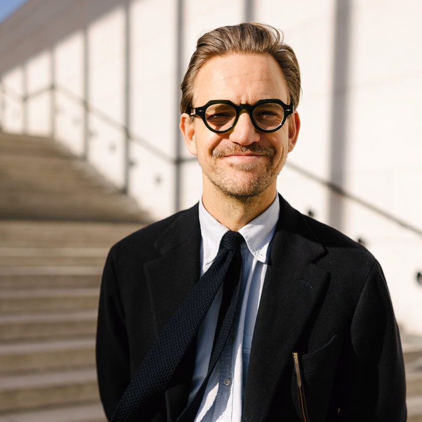 Sebastian Tigges mit runder Brille, dunklem Sakko und Strickkrawatte steht lächelnd vor einer hellen Steintreppe.