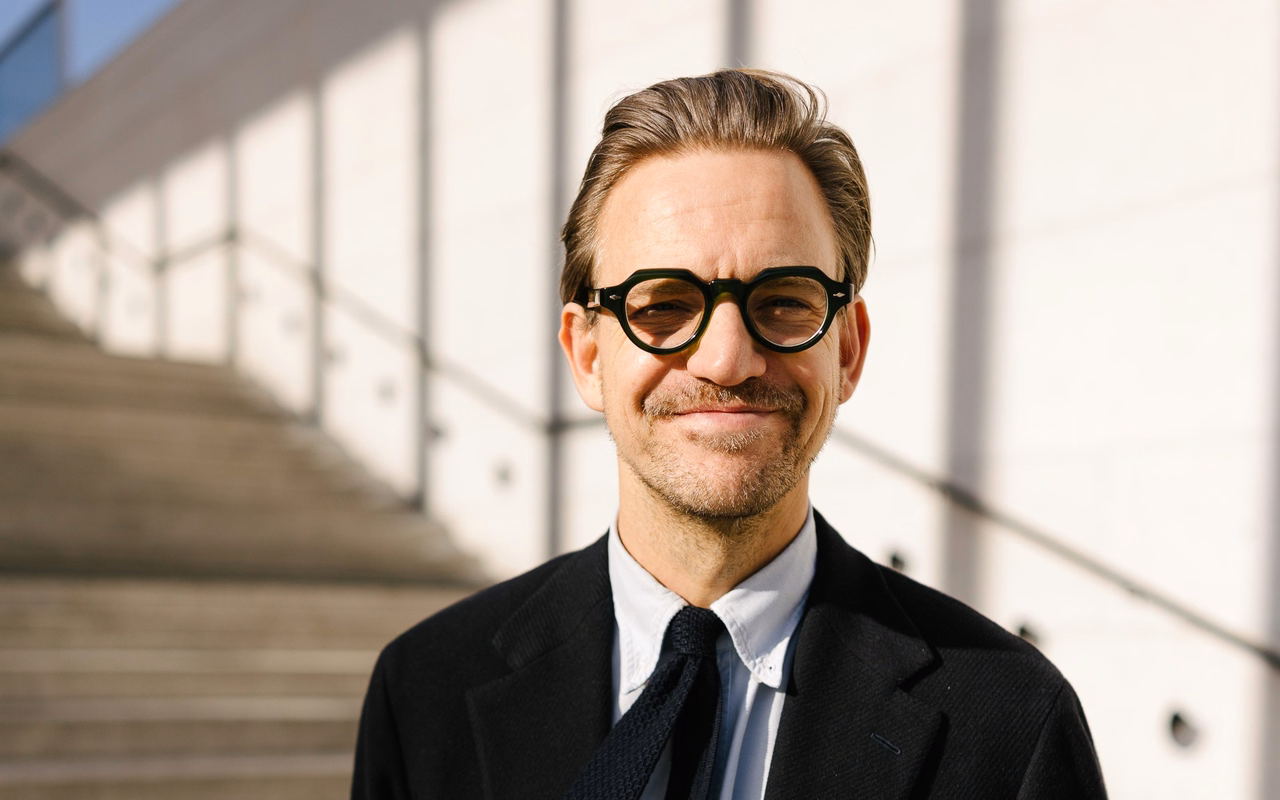Nahaufnahme von Sebastian Tigges mit runder Brille und Strickkrawatte, lächelnd vor einer sonnigen Steintreppe.
