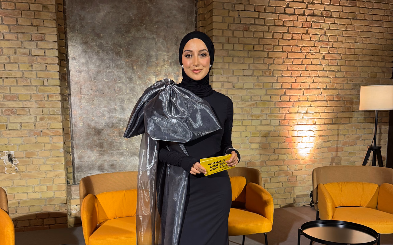 Büsra Sayed in schwarzem Kleid mit silberner Schleife vor einer Backsteinwand zwischen gelben Sesseln, in der Hand eine Karte des Women's Business Iftar 2026.