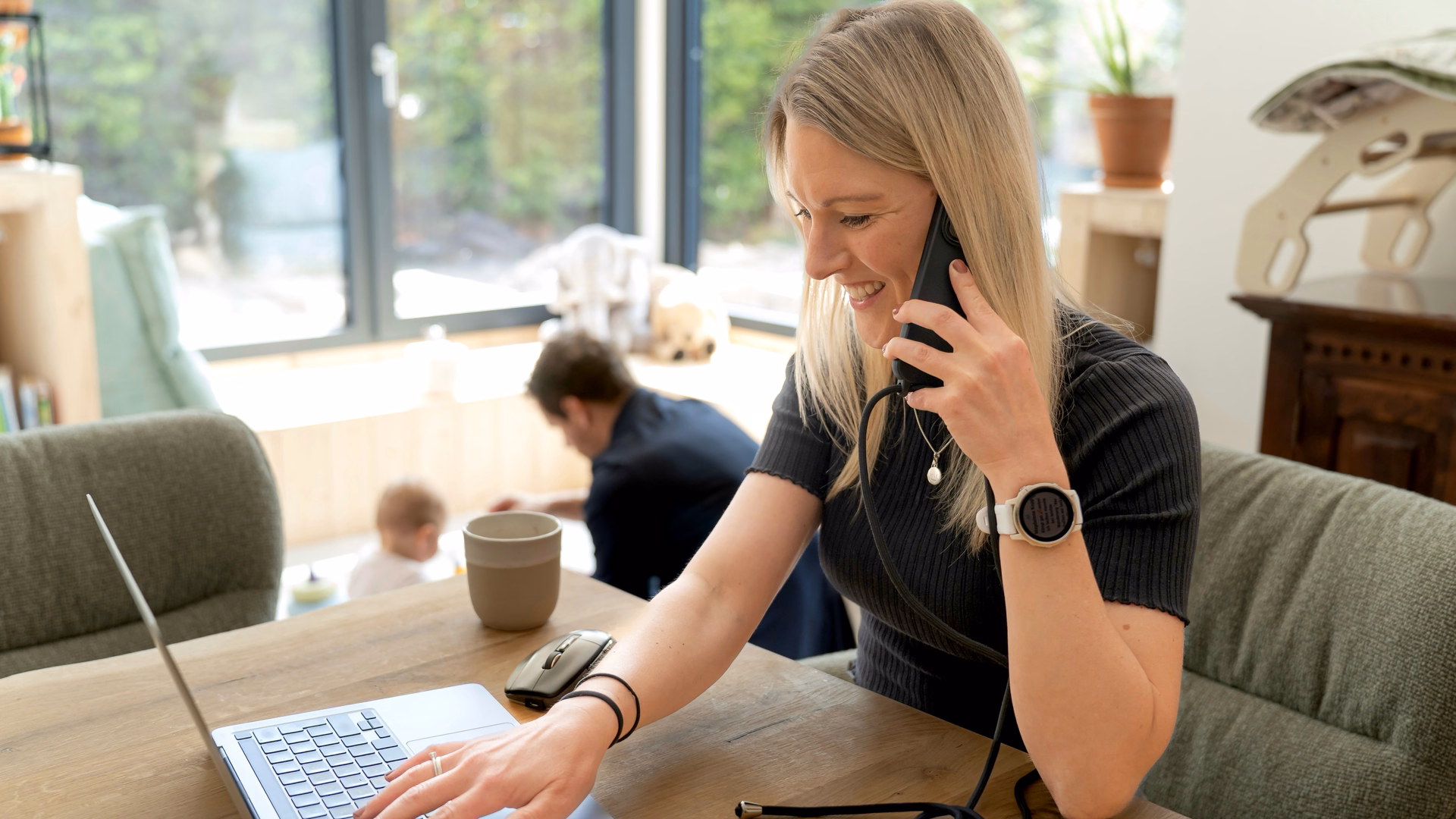 Hanna Wagner telefoniert lächelnd am Esstisch und tippt auf ihrem Laptop, im Hintergrund spielt ihre Tochter.