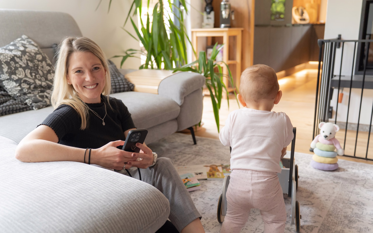 Hanna Wagner sitzt mit dem Handy vor dem Sofa, ihre Tochter schiebt einen Lauflernwagen durchs Wohnzimmer.
