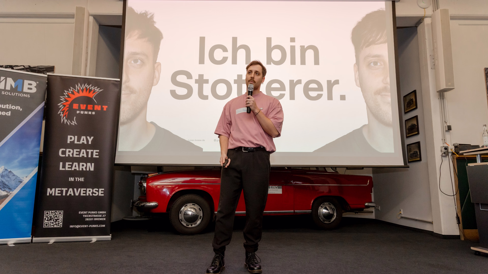 Lucas Böse steht mit Mikrofon auf einer Bühne, hinter ihm eine Leinwand mit dem Satz „Ich bin Stotterer.“ und davor ein roter Oldtimer.