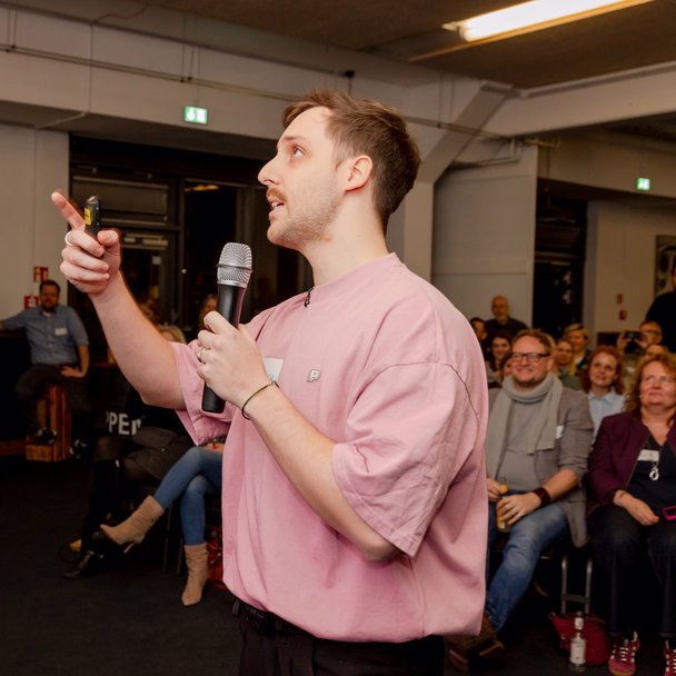Lucas Böse zeigt mit dem Mikrofon in der Hand nach oben, rechts von ihm sitzt das Publikum.