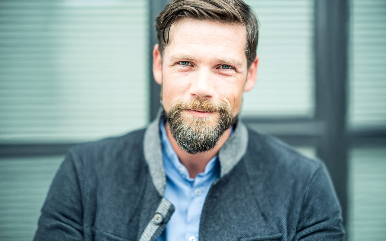 Thomas Hann mit Vollbart, grauer Jacke und hellblauem Hemd lächelt vor einer Fensterfront in die Kamera.