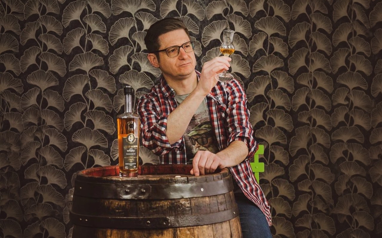 Mathias Gerstner hält ein Glas Whisky ins Licht, vor ihm auf einem Holzfass steht eine Flasche Whisky.