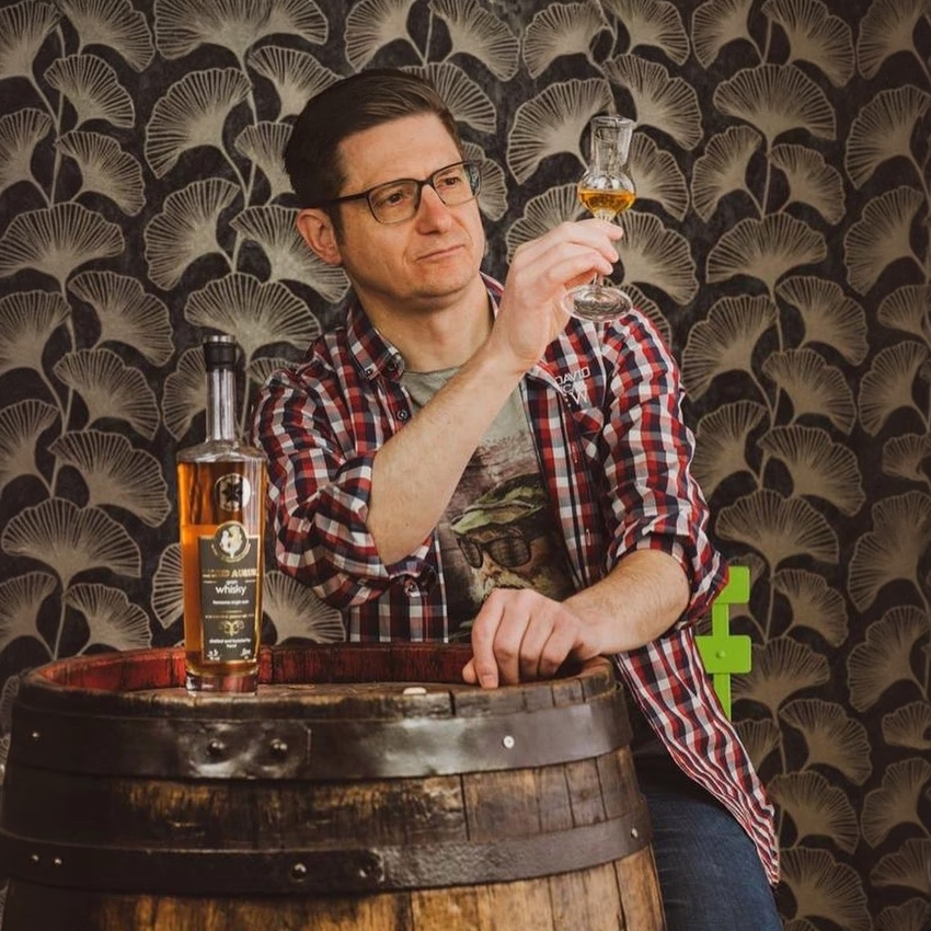 Mathias Gerstner lehnt an einem Holzfass und betrachtet ein Glas Whisky, daneben steht eine Flasche.