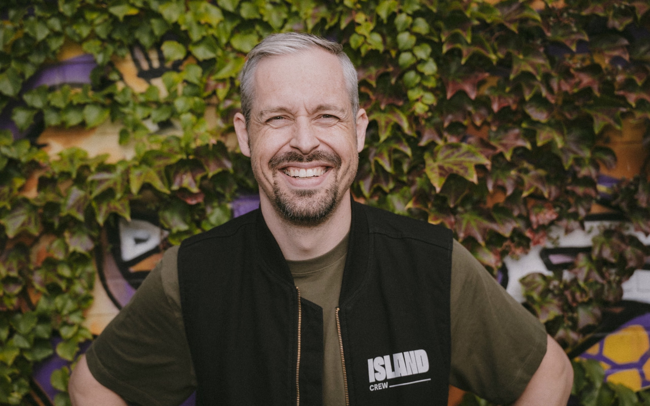 Florian Laudon lacht in die Kamera, er trägt eine schwarze Weste mit Island-Crew-Aufdruck vor einer efeubewachsenen Graffiti-Wand.