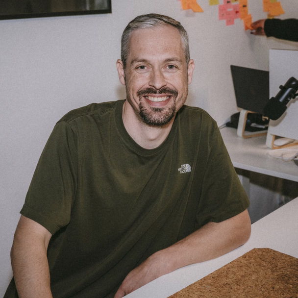 Florian Laudon sitzt lächelnd im olivgrünen T-Shirt an einem Schreibtisch, hinter ihm hängen bunte Haftnotizen an der Wand.