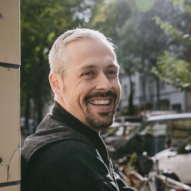 Florian Laudon lehnt lachend an einer Säule, hinter ihm liegt eine sonnige Straße mit Bäumen und geparkten Fahrrädern.