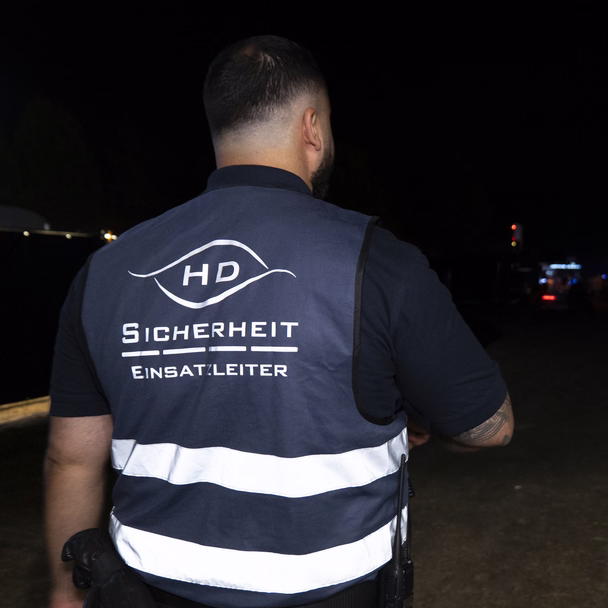 Ein Einsatzleiter von HD Sicherheit in Warnweste steht nachts vor einer beleuchteten Veranstaltungsbühne, von hinten fotografiert.