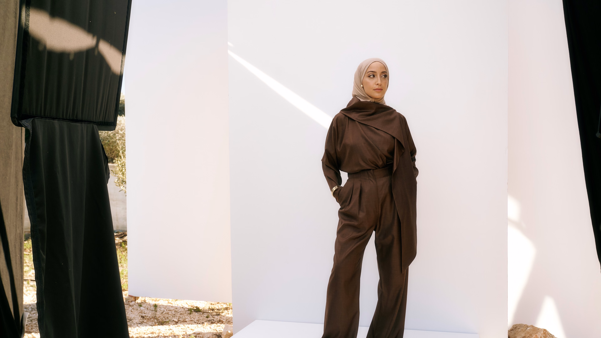 Büsra Sayed in braunem Hosenanzug und hellem Hijab vor weißen Stellwänden bei einem Fotoshooting im Freien.