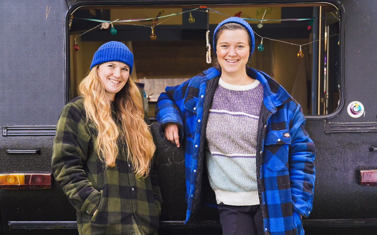 Sophie Rönnau und Christina Rüschhoff stehen lachend in Karojacken und blauen Mützen vor einem schwarzen Wohnwagen mit Lichterkette.