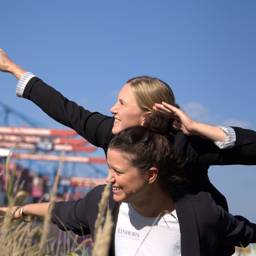 Sophie Rönnau und Christina Rüschhoff breiten huckepack die Arme aus wie Flügel, dahinter stehen Containerbrücken am Hafen.